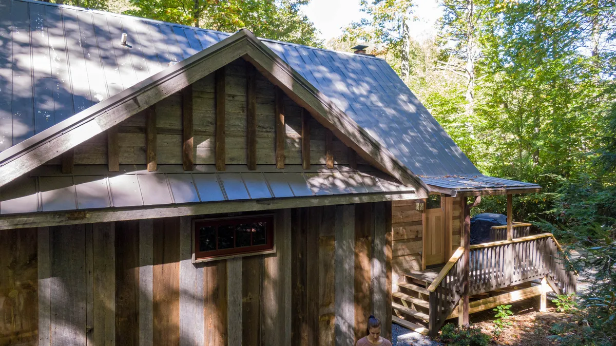 This exterior image features a charming wooden structure with a metal roof nestled among lush greenery. The inviting front porch provides a clear connection to the outdoors, with comfortable seating and scenic natural surroundings that hint at year-round enjoyment, relaxation, and nature appreciation.