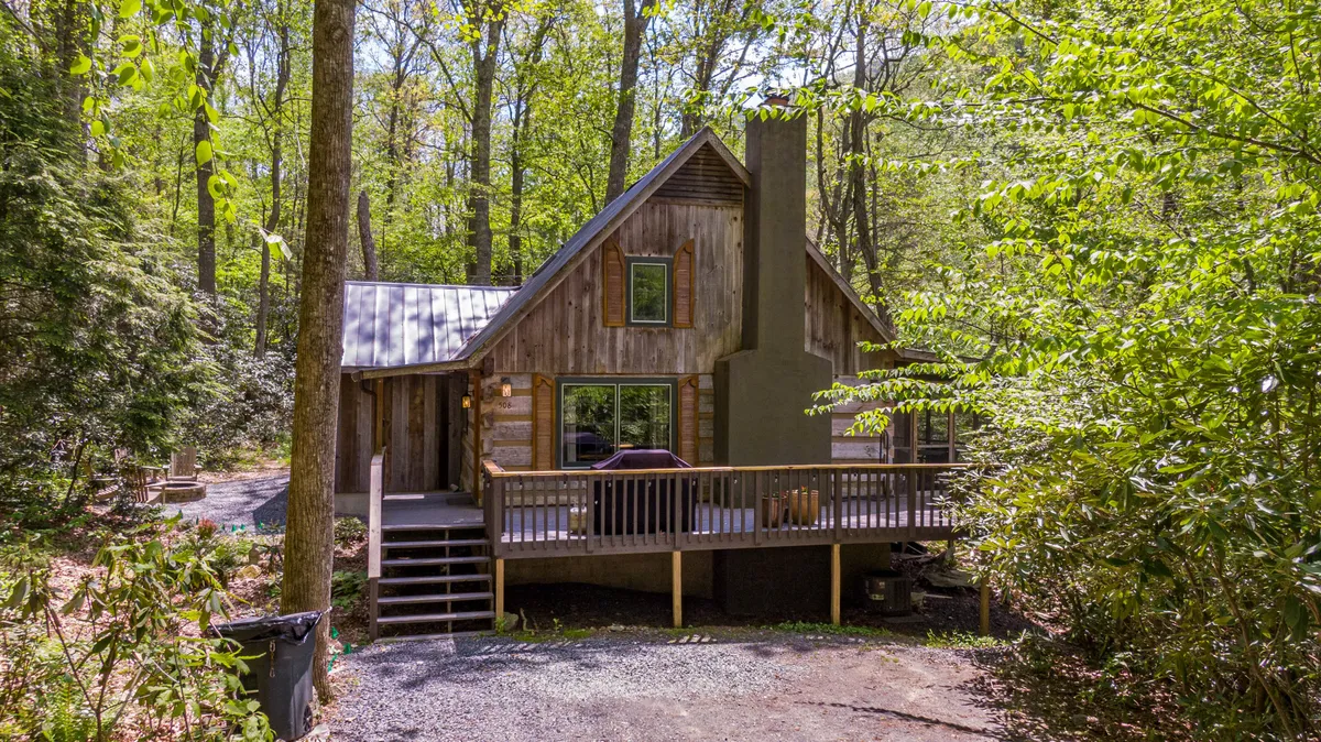 A wide exterior view of the wooden cabin with a large deck nestled in abundant greenery. The scene shows the deck as a comfortable transition space between the home and the wild outdoors, perfect for relaxing or gathering before or after adventures.