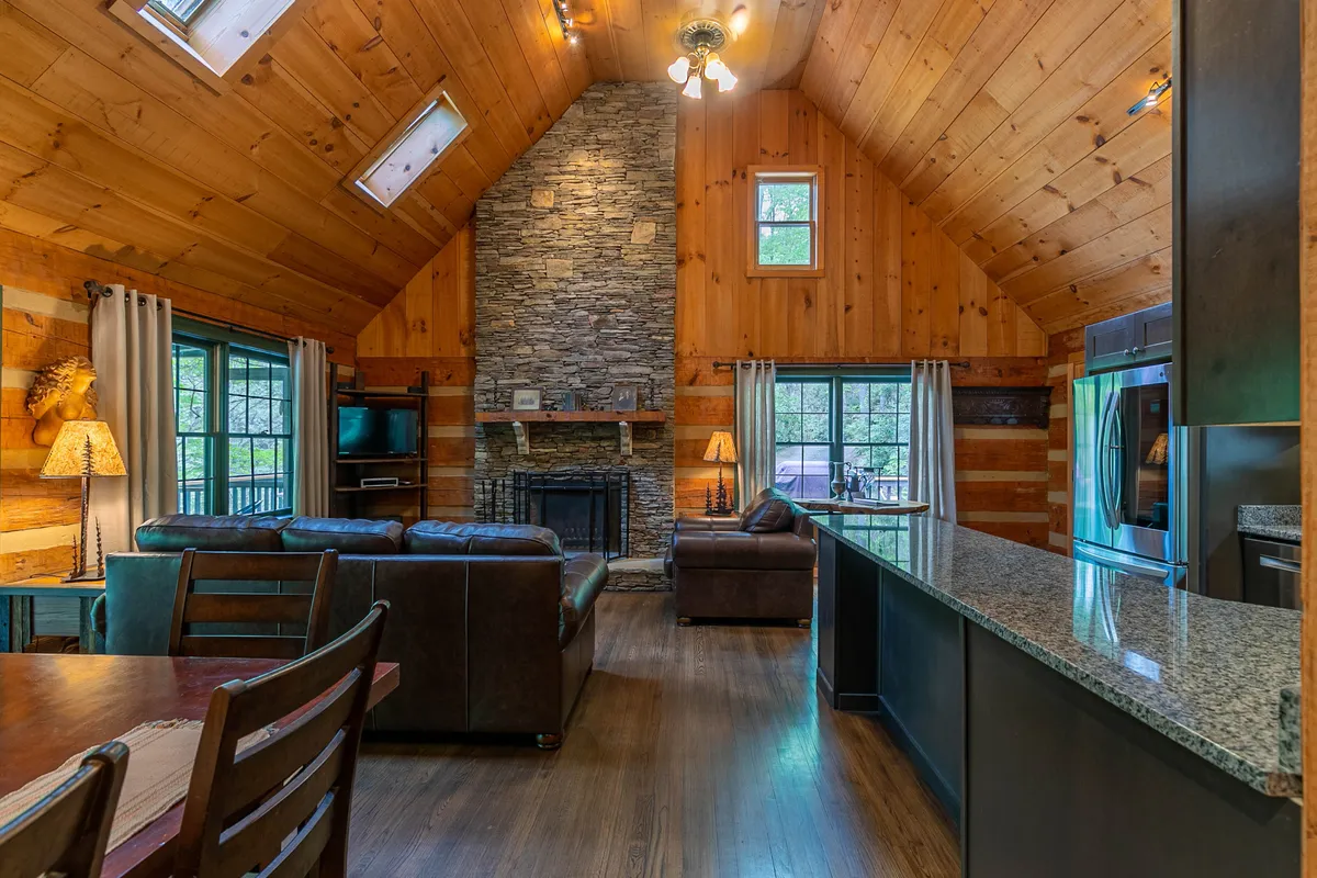 An expansive living and dining area with a stone fireplace, wooden beams, large windows that bring in natural light, a leather sofa, and a dining table. The setting is perfect for family gatherings, cozy evenings, and shared meals, with views that suggest a connection to the outdoors.