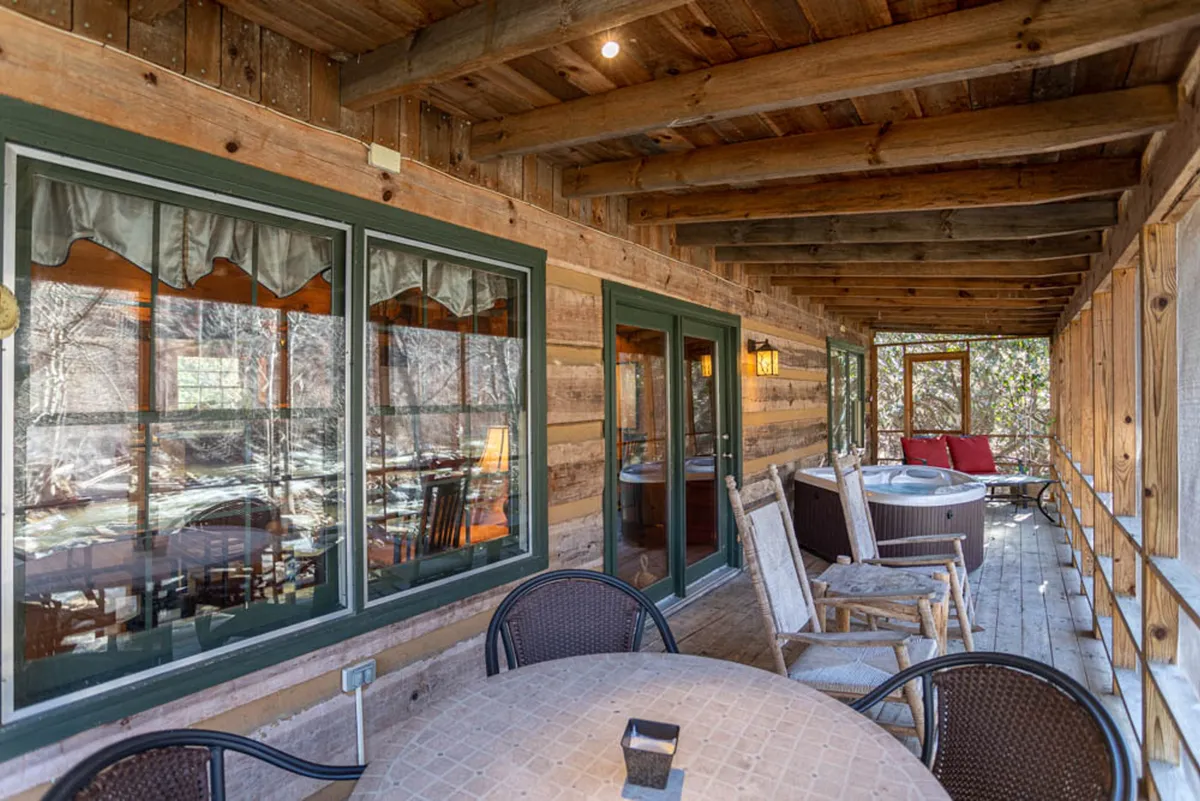 This image features an inviting wooden deck framed by large windows, a hot tub, and an outdoor seating area. The deck is surrounded by natural greenery, creating a tranquil, secluded atmosphere perfect for relaxation and connection with nature.