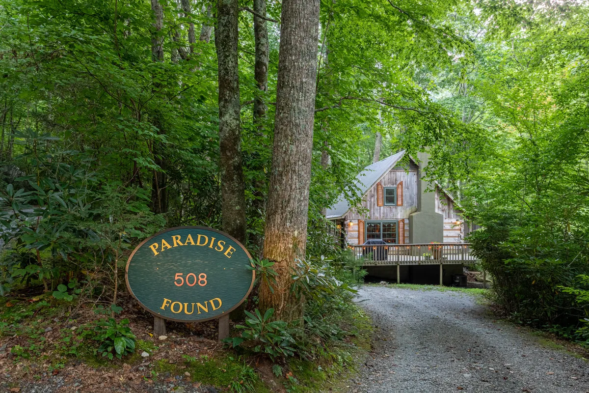The image showcases a charming wooden cabin nestled among lush trees, with a welcoming sign reading 'PARADISE FOUND' prominently displayed. The tranquil forest setting surrounds the cabin, hinting at privacy and a deep connection with nature. The scene radiates serenity, inviting guests to imagine peaceful mornings on the porch and cozy evenings by the fire.
