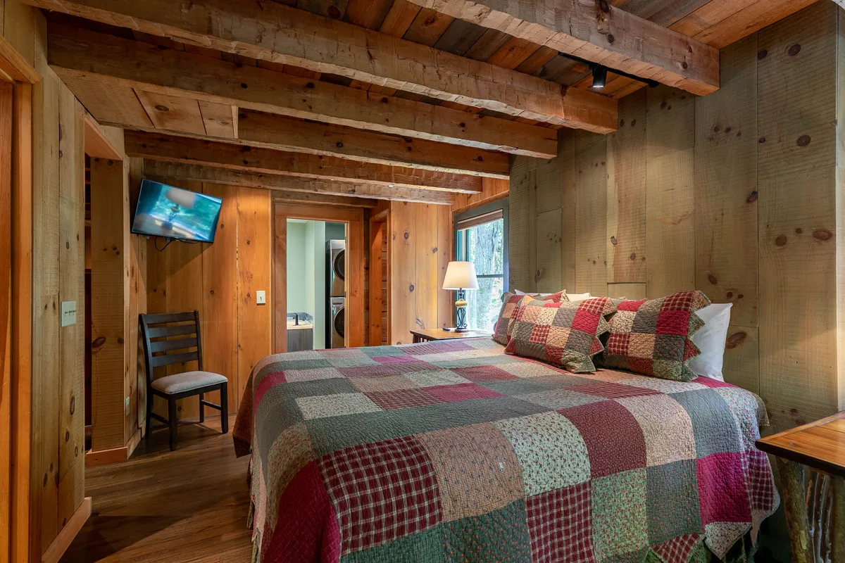 A master bedroom retreat with a hand-hewn beam ceiling, a king bed dressed in a quilted bedspread, a large window that brings in morning light, and a TV for private relaxation. The room feels both inviting and luxurious, perfect for intimate nights in.