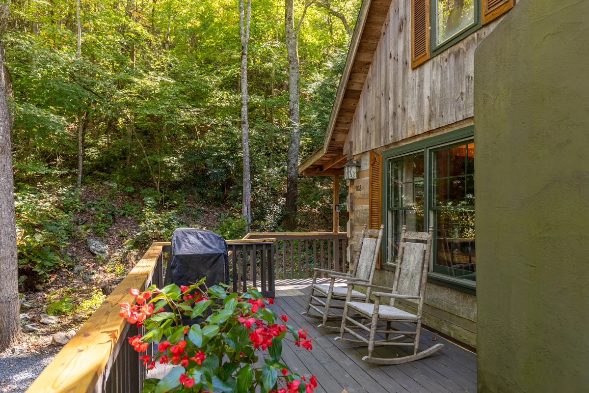 A spacious wooden deck surrounded by trees, featuring rocking chairs, a flower pot, and a grill. The setup invites guests to relax, socialize, enjoy morning coffee, or barbecue as the sun sets. The lush greenery offers privacy and a strong connection to nature.
