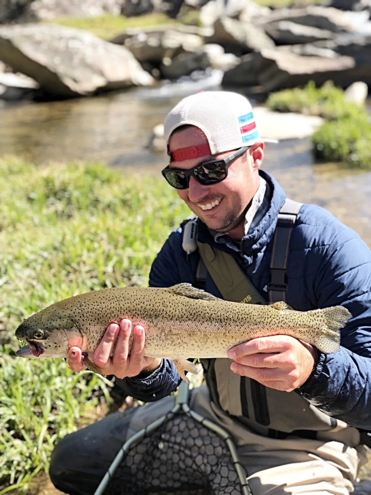 A person proudly holds a large trout on the bank of a clear, rocky river bordered by grass, embodying the excitement of catch-and-release fishing. The surrounding nature further emphasizes the property’s connection to outdoor pursuits.