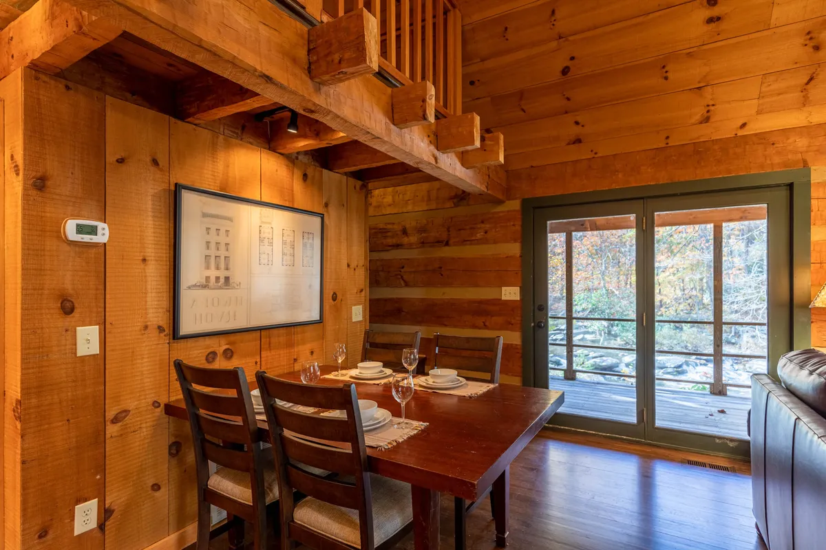 A dining area with a wooden table, rustic wooden walls, and large windows providing ample natural light. The space is inviting for family meals, game nights, and relaxed conversations, embodying the warmth and tradition of repeated visits.