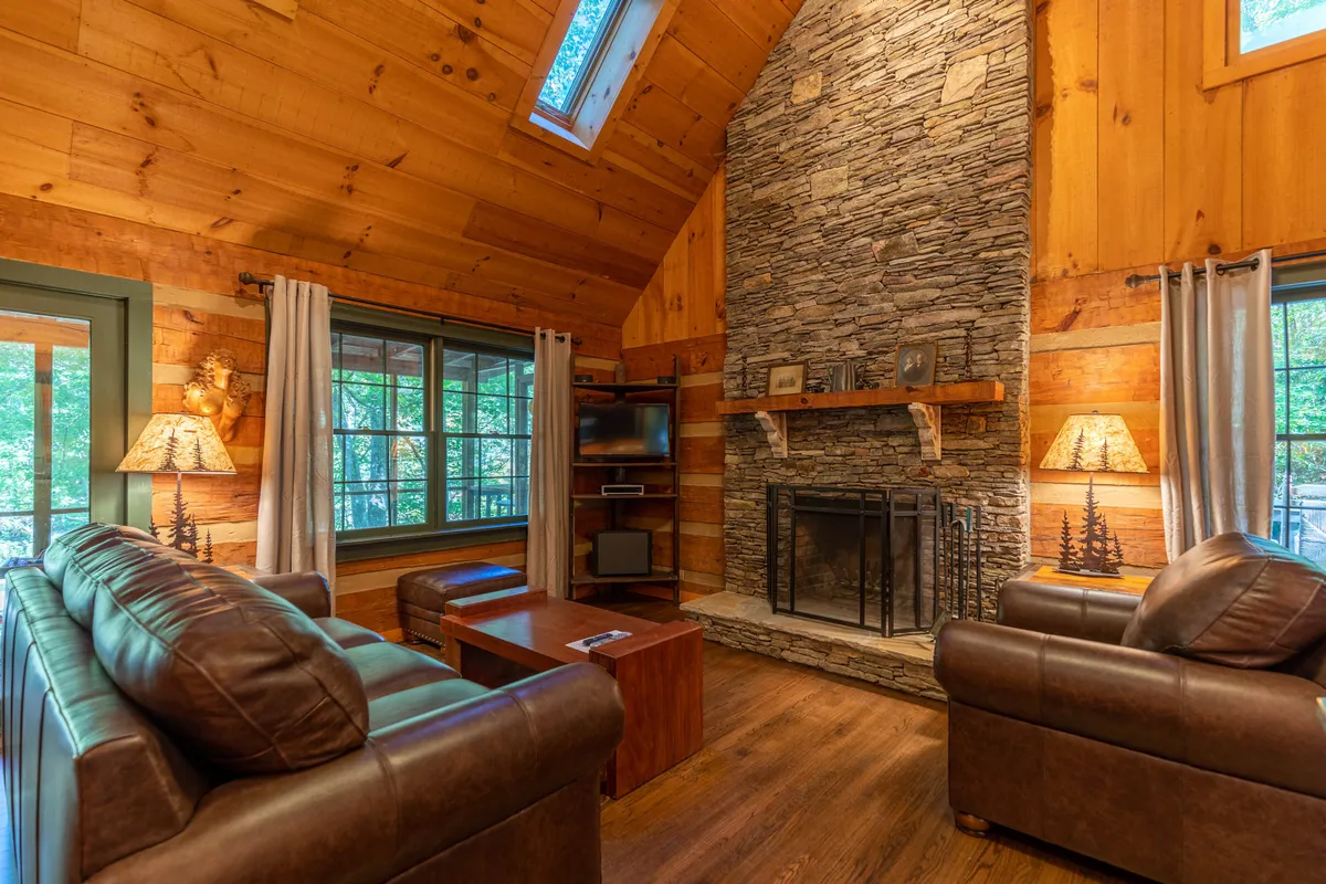 A warm, welcoming living room with a large stone fireplace as the focal point, wooden beams overhead, comfortable leather sofas, and expansive windows that let in natural light. The setup suggests cozy relaxation and socializing, and the window hints at partial river views.