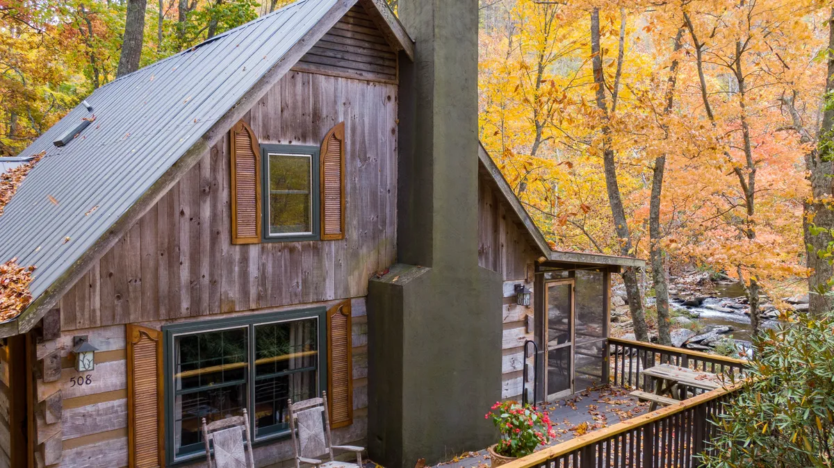 A rustic wooden cabin sits amid colorful autumn foliage, with a deck furnished for relaxation and surrounded by tranquil woods. The image highlights the property’s scenic grounds and seasonal beauty.
